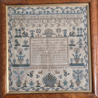 A framed antique cross-stitch sampler with blue embroidery on cream fabric, featuring a central text panel, floral motifs, and a decorative border, enclosed in a brown wooden frame.